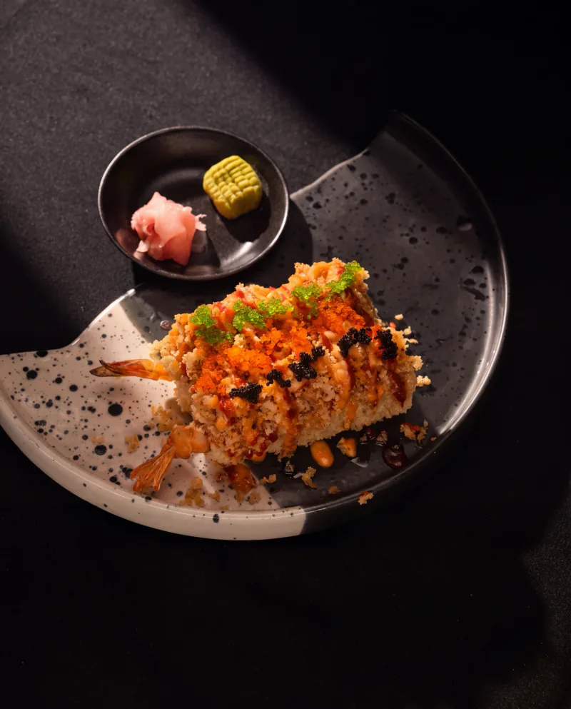 A crunchy prawn roll topped with tobiko, with pickled ginger and wasabi on a speckled plate